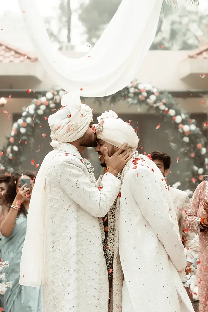 Two grooms in white wedding attire share a kiss.
