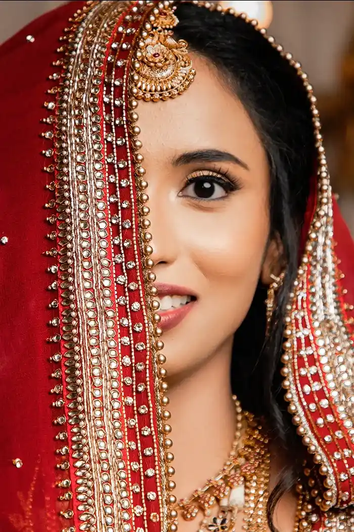 Half of a smiling bride’s face is visible beneath an ornate red veil adorned with gold embroidery